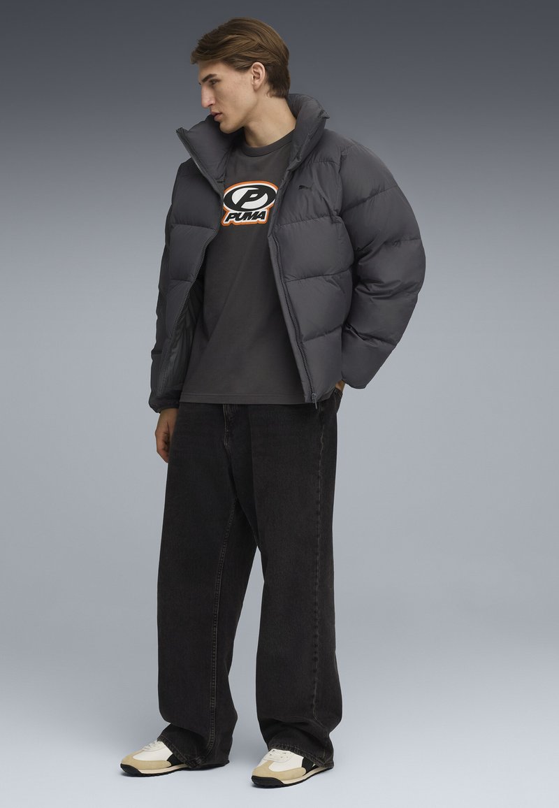 Grijze pufferjack met een hoge kraag, gedragen over een donker T-shirt met een logo. Gecombineerd met losse zwarte jeans en sneakers met beige accenten.