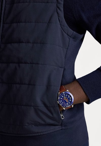 Asmuo dėvi tamsiai mėlyną susegamą liemenę ir megztinį, ranka kišenėje, demonstruojantis laikrodį su mėlynu ciferblatu ir ruda odine apyranke.