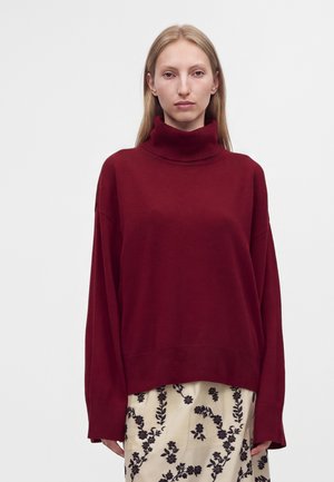 Femme aux cheveux blonds et raides portant un pull à col roulé ample rouge foncé et une jupe crème avec des motifs floraux noirs sur un fond uni.