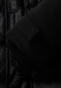 Cuff della giacca imbottita nera caratterizzato da una texture morbida con chiusura in Velcro. Il tessuto è liscio con dettagli trapuntati visibili sul retro.