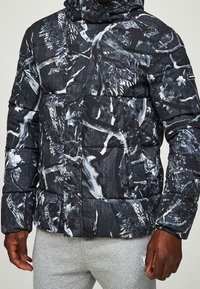 Muž oblečený v pufferové bundě s kapucí v černé a šedé kamufláž vzoru a světle šedých teplácích, stojící před jednobarevným pozadím.