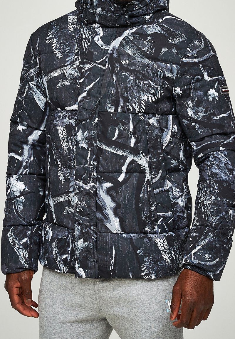 Muž oblečený v pufferové bundě s kapucí v černé a šedé kamufláž vzoru a světle šedých teplácích, stojící před jednobarevným pozadím.