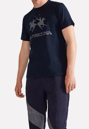 Homme portant un t-shirt bleu marine à manches courtes avec un logo de joueurs de polo croisés et du texte, associé à un pantalon foncé avec des panneaux diagonaux gris.