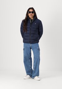 Jachetă puffer albastră cu fermoar frontal și insigne, asortată cu blugi cargo casual într-o nuanță deschisă de albastru și adidași albi. Modelul poartă ochelari de soare.