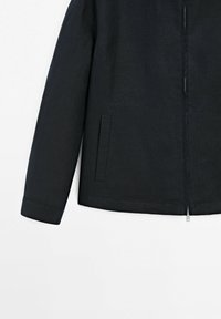 Veste noire à manches longues, fermeture éclair frontale et une poche verticale fendue sur le côté gauche, présentée sur fond blanc.