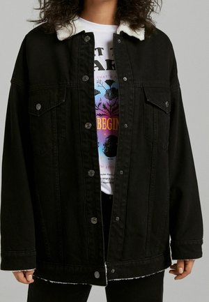 Osoba w rozpiętej czarnej kurtce dżinsowej z białym kołnierzem z futerka nosi białą koszulkę z kolorowym kwiatowym wzorem i napisem.