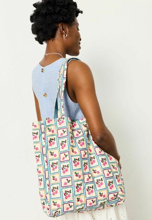 Frau mit kurzen lockigen Haaren trägt eine große Tragetasche mit einem bunten Muster aus Radieschen in eingerahmten Quadraten.