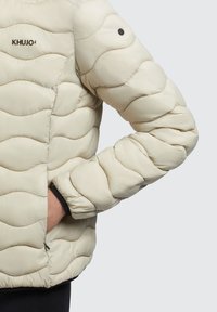 Jachetă pufosă de culoare crem, cu un model ondulat de matlasare, având un logo negru și un buzunar cu fermoar pe lateral. Mansetele au un accent negru.