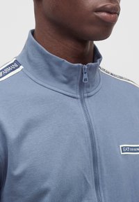 Крупен план на човек, носещ синя ципна яке EA7 Emporio Armani с лого пачове на гърдите и раменната ивица.