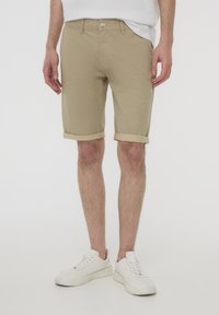 Pantaloni corti beige realizzati in tessuto texturizzato, con orli arrotolati e chiusura con bottone. Indossati con scarpe da ginnastica bianche, appoggiati su uno sfondo neutro.