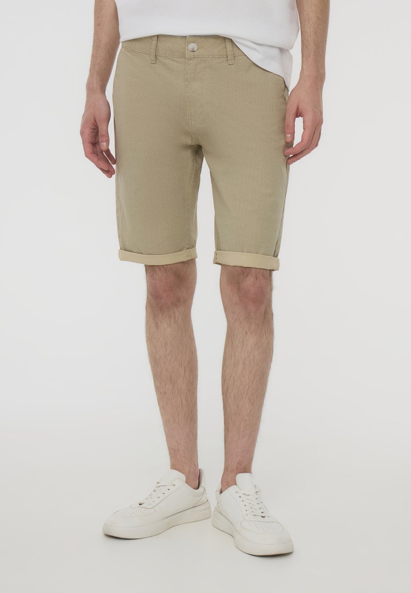 Pantaloni corti beige realizzati in tessuto texturizzato, con orli arrotolati e chiusura con bottone. Indossati con scarpe da ginnastica bianche, appoggiati su uno sfondo neutro.