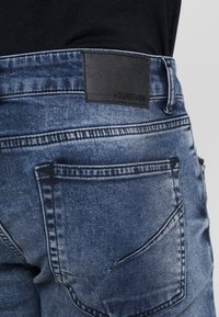 Närbild av ljusblå denimjeans bakficka med svart läderpatch och synlig sömnad på person som bär svart topp.