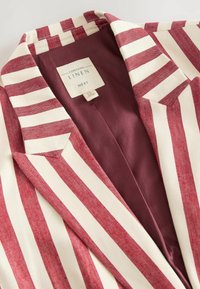 Blazer en lin avec des rayures verticales rouges et blanches, col à revers, et doublure bordeaux avec une étiquette indiquant la composition du matériau.