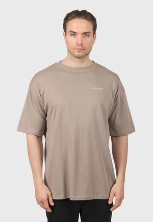 Homme aux cheveux gominés portant un t-shirt beige ample avec le texte "KLEINIGKEIT" sur la poitrine, se tenant devant un fond clair uni.