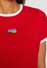 Camiseta roja de algodón, con un ribete blanco en el cuello redondo. Incluye un logo azul y amarillo "HILFIGER 1985 NYC U.S.A" en el pecho.
