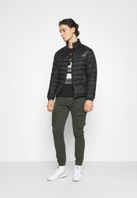 Zwarte pufferjack met een hoge kraag, gequilt textuur; gecombineerd met olijfgroene cargo broek en witte sneakers. Eenvoudig grafisch T-shirt eronder.