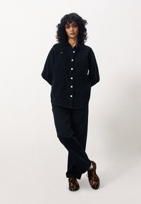 Chemise en velours côtelé noir avec deux poches poitrine et boutons blancs, associée à un pantalon noir et des chaussures marron à imprimé léopard. Texture douce tout au long.