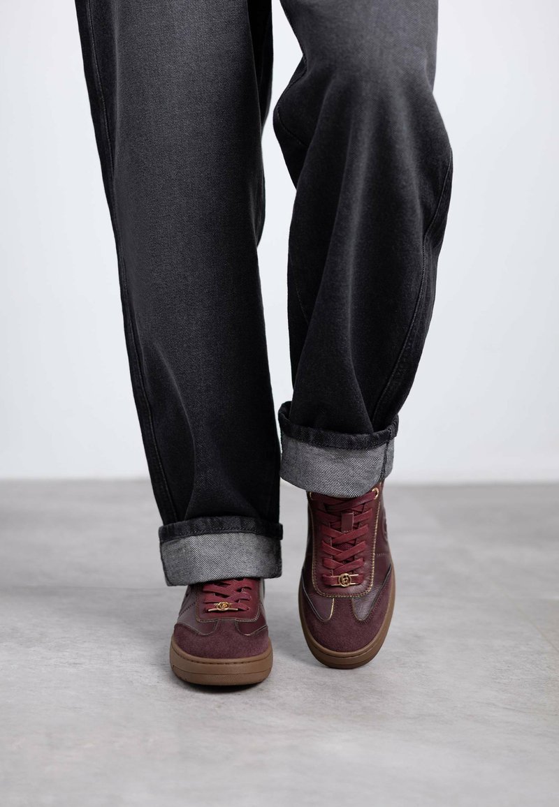 Zapatillas marrón con paneles de ante y cuero, que cuentan con suelas de goma marrón y pantalones de mezclilla gris arremangados. La textura es suave con colores contrastantes.