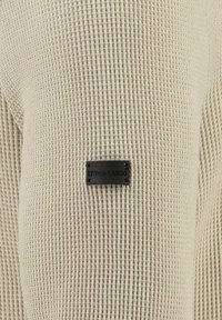Maglione lavorato a maglia beige con motivo testurizzato, dotato di un piccolo etichetta nera con la scritta "KEY LARGO" vicino al centro.