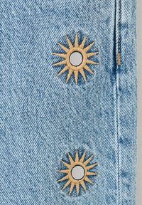 Tissu en denim bleu clair avec deux écussons brodés en forme de soleil, chacun orné d'un bouton métallique au centre, cousus près d'une couture verticale.