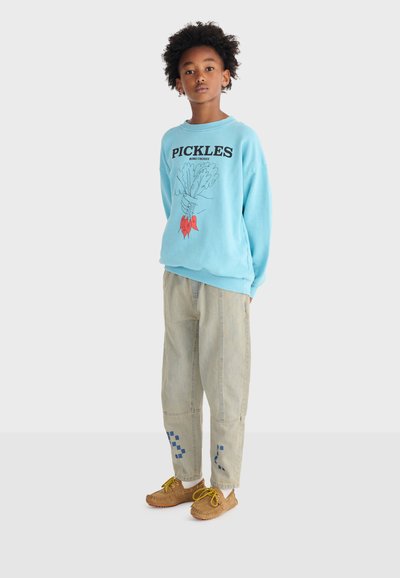 Enfant aux cheveux bouclés portant un sweat-shirt bleu "PICKLES", un pantalon beige avec des motifs carrés bleus, et des mocassins marron avec des lacets jaunes.