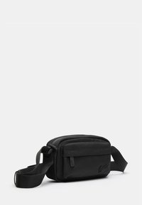 Crni platneni crossbody ruksak s zatvaračem, pravokutnog oblika, podesivim remenom i reljefnim logotipom sprijeda.
