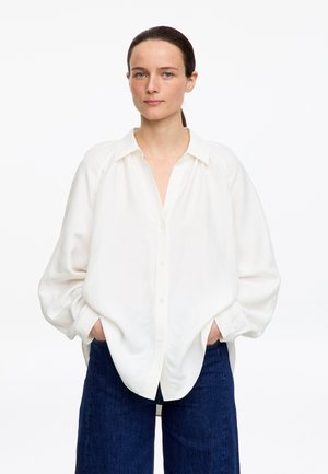 Femme aux cheveux bruns attachés, portant une chemise blanche ample à boutons et un pantalon bleu foncé, debout les mains dans les poches contre un fond blanc.