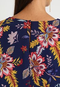 Tissu à motif floral bleu marine avec de grandes fleurs roses et jaunes, featuring une ouverture arrière fendue. Texture lisse aux couleurs vives.