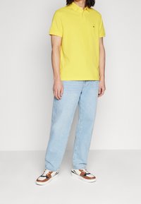 Keltainen polo-paita, vaaleansiniset löysät farkut ja ruskeat sekä valkoiset tennarit. Polossa on pieni logo rinnassa; rento asuste.