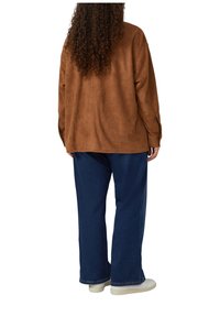 Person mit langen, lockigen Haaren, der eine braune Wildlederjacke, blaue Schlagjeans und weiße Sportschuhe trägt, steht mit dem Rücken zur Kamera.