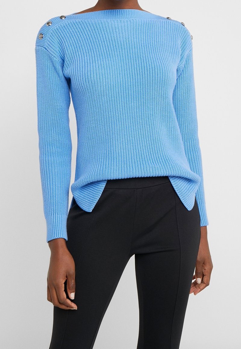 Femme portant un pull en maille bleu clair avec des boutons aux épaules et un pantalon noir ajusté sur un fond uni.