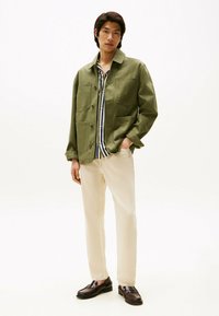 Olivgrüne Jacke mit zwei vorderen Taschen und spitzem Kragen, über ein gestreiftes Hemd geschichtet, kombiniert mit cremefarbenen Hosen und dunklen Schuhen.