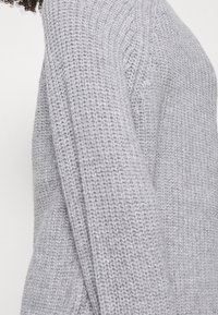 Šedý pletený svetr se zřetelným vzorem, volným střihem a žebrovanými detaily podél výstřihu a rukávů.