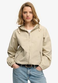 Chaqueta beige con cremallera y capucha, puños elásticos y detalles de cordón. Cuenta con bolsillos laterales y un logo discreto en el pecho. Corte casual.