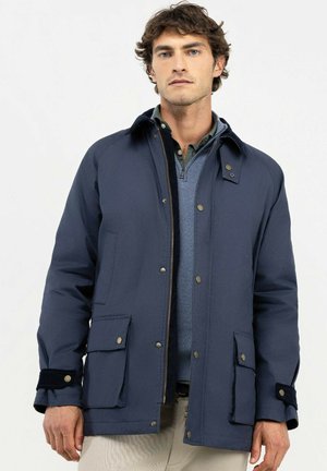 Hombre con chaqueta azul marino sobre un suéter azul con cremallera y camisa verde, de pie contra un fondo claro y liso.