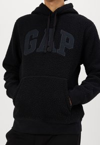 Persona che indossa una felpa nera pelosa con la scritta "GAP" sul petto, con le mani nella tasca frontale, su uno sfondo semplice.