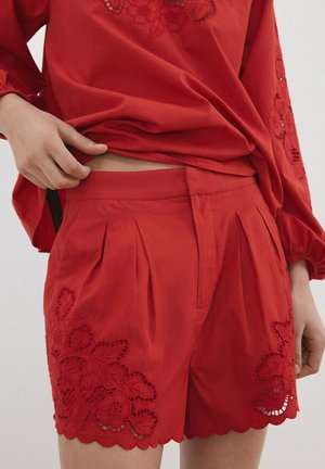 Pantalones cortos rojos con bordados florales de encaje y dobladillos en pico, usados con una parte superior de manga larga bordada a juego, ajustando la cintura con la mano.