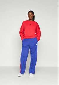 Raudonas per didelis džemperis su juodu Adidas logotipu; mėlyni treniruočių kelniai su šoninėmis juostelėmis ir logotipu. Dėvimi su baltais batais. Vienodas fonas.