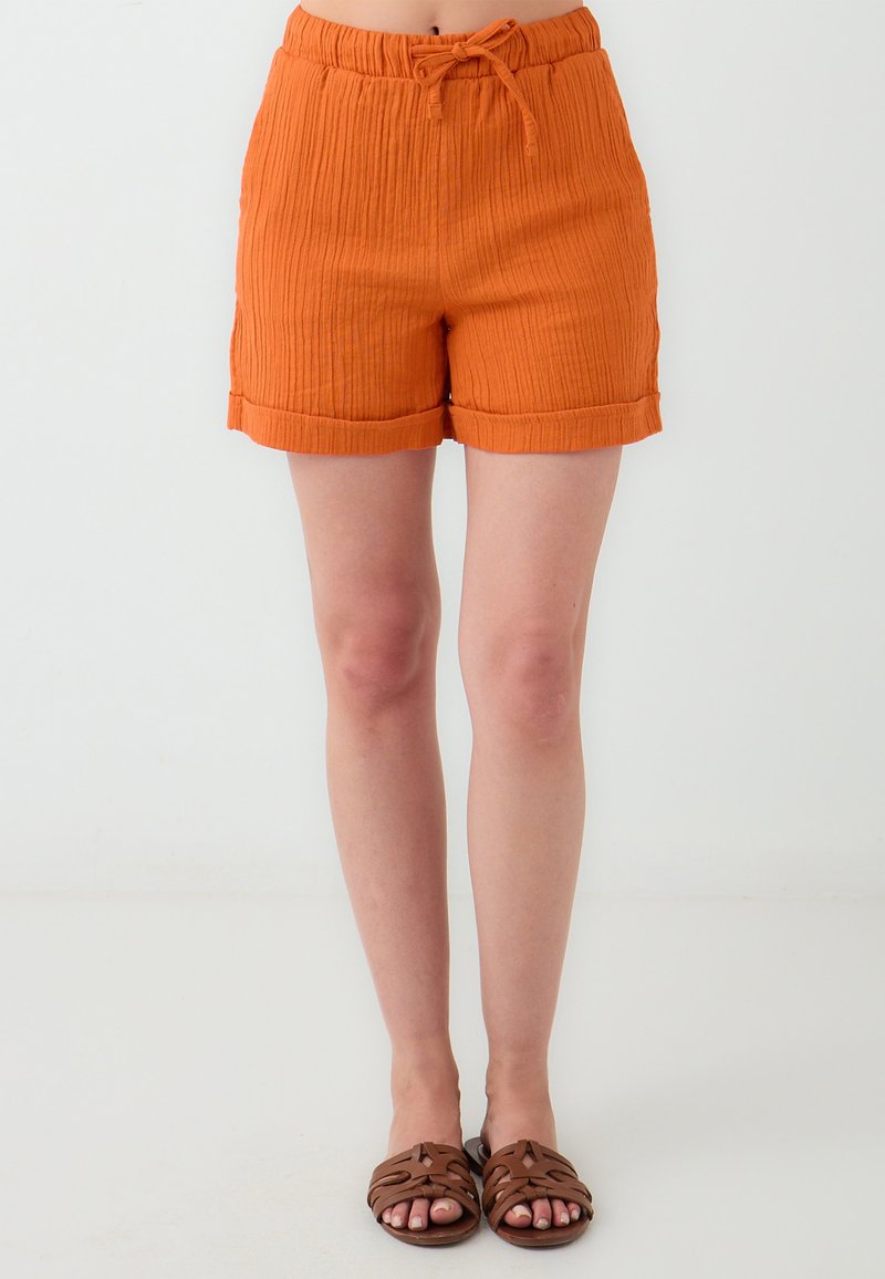 Oranje geribde shorts met een trekkoord in de taille, omgerolde zomen en een relaxte pasvorm, gecombineerd met bruine sandalen met banden op blote benen.