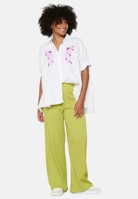 Bílá oversized košile s růžovou květinovou výšivkou, vynikající s světle zelenými, texturovanými širokými kalhotami. Model nosí bílé tenisky.