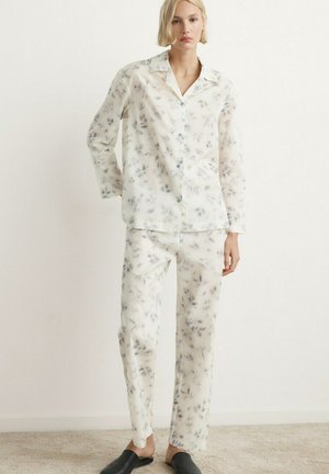 Donna che indossa un completo pigiama in seta bianca con stampa floreale grigia e scarpe nere senza lacci, in piedi davanti a una parete bianca liscia.