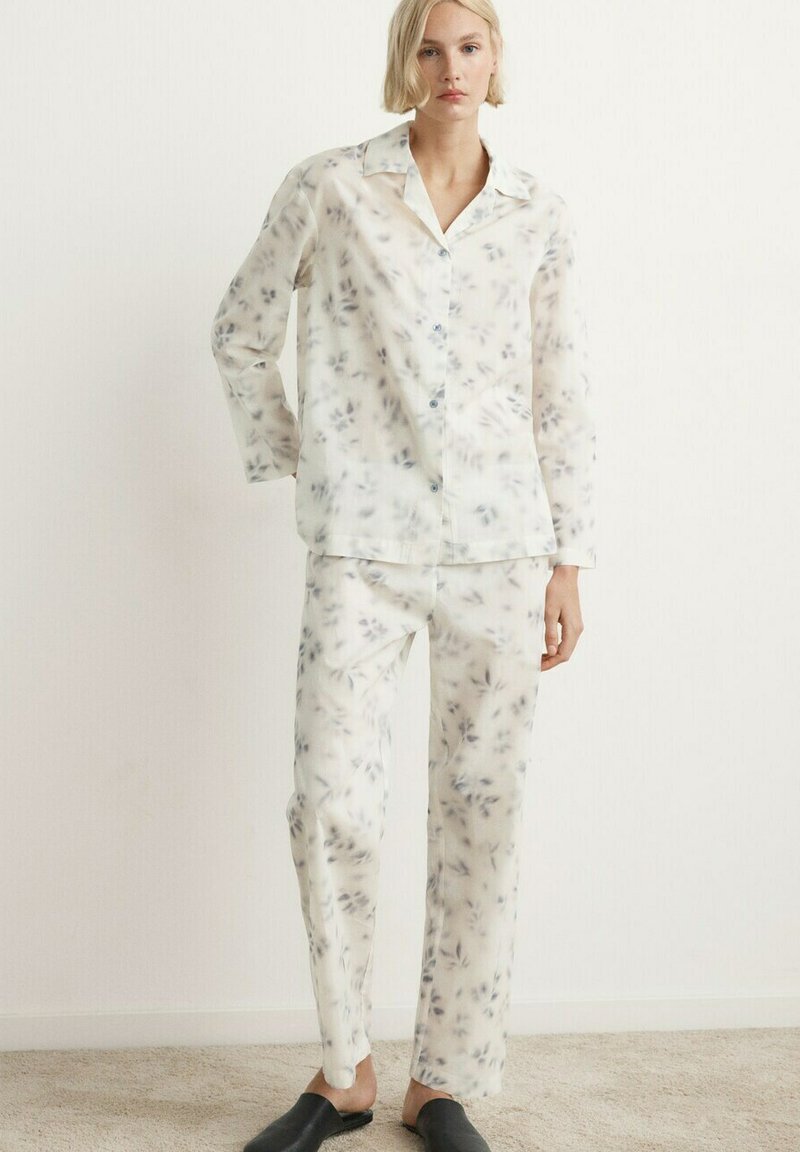 Femme portant un ensemble de pyjama en soie blanche avec un imprimé floral gris et des chaussures à enfiler noires, debout contre un mur blanc uni.