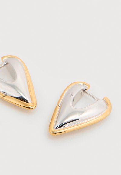Ασημένια και χρυσά σκουλαρίκια με περίγραμμα σε σχήμα τόξου με αιχμηρές άκρες και λεία, γυαλιστερή επιφάνεια, τοποθετημένα σε ανοιχτό φόντο.