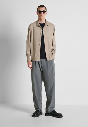 Man draagt zwarte zonnebril, beige overhemd met knopen, zwart t-shirt, losse grijze broek en zwarte schoenen, staand tegen een effen lichte achtergrond.