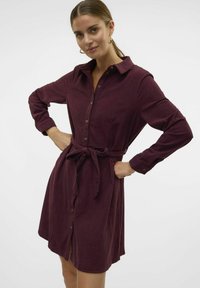 Robe boutonnée bordeaux à manches longues et taille liée, en tissu texturé. Présente un col et une fente à l'avant.