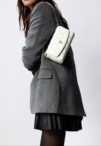 Sac en cuir blanc matelassé avec quincaillerie dorée, présentant une fermeture à rabat et une anse en chaîne, porté contre une veste grise.