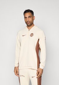 Crèmekleurige hoodie met bruine accenten en zijstrepen, voorzien van een ritskraag en een logo op de borst. Soepele stofstructuur.