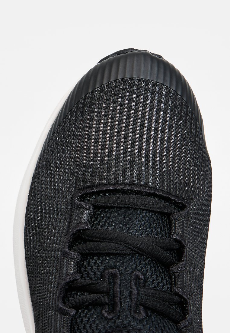 Zapatilla deportiva negra con parte superior de malla texturizada, punta acanalada y cordones negros. Presenta una suela blanca lisa.