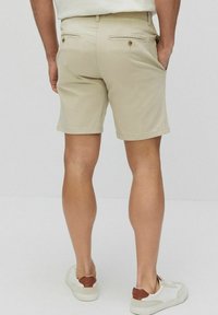 Beige shorts i en slät текстur, bakfickor med guldknappar och en ren fåll. Bärs med vita sneakers med bruna detaljer.