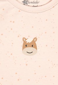 Helles rosa Baumwoll-T-Shirt mit einem lächelnden Pferdekopf-Design in Braun- und Weißtönen, umgeben von kleinen, verstreuten orangen Punkten.